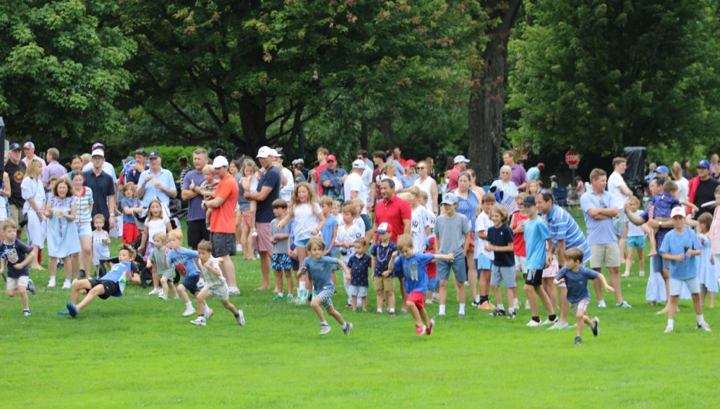 A group of people participating in a community event at a park, with tents set up, children playing, and trees in the background.