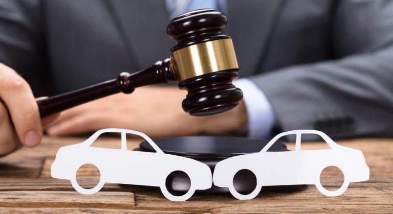 A car accident lawyer standing confidently with arms crossed, wearing a suit, in front of a blurred image of a damaged car and emergency responders at the scene.